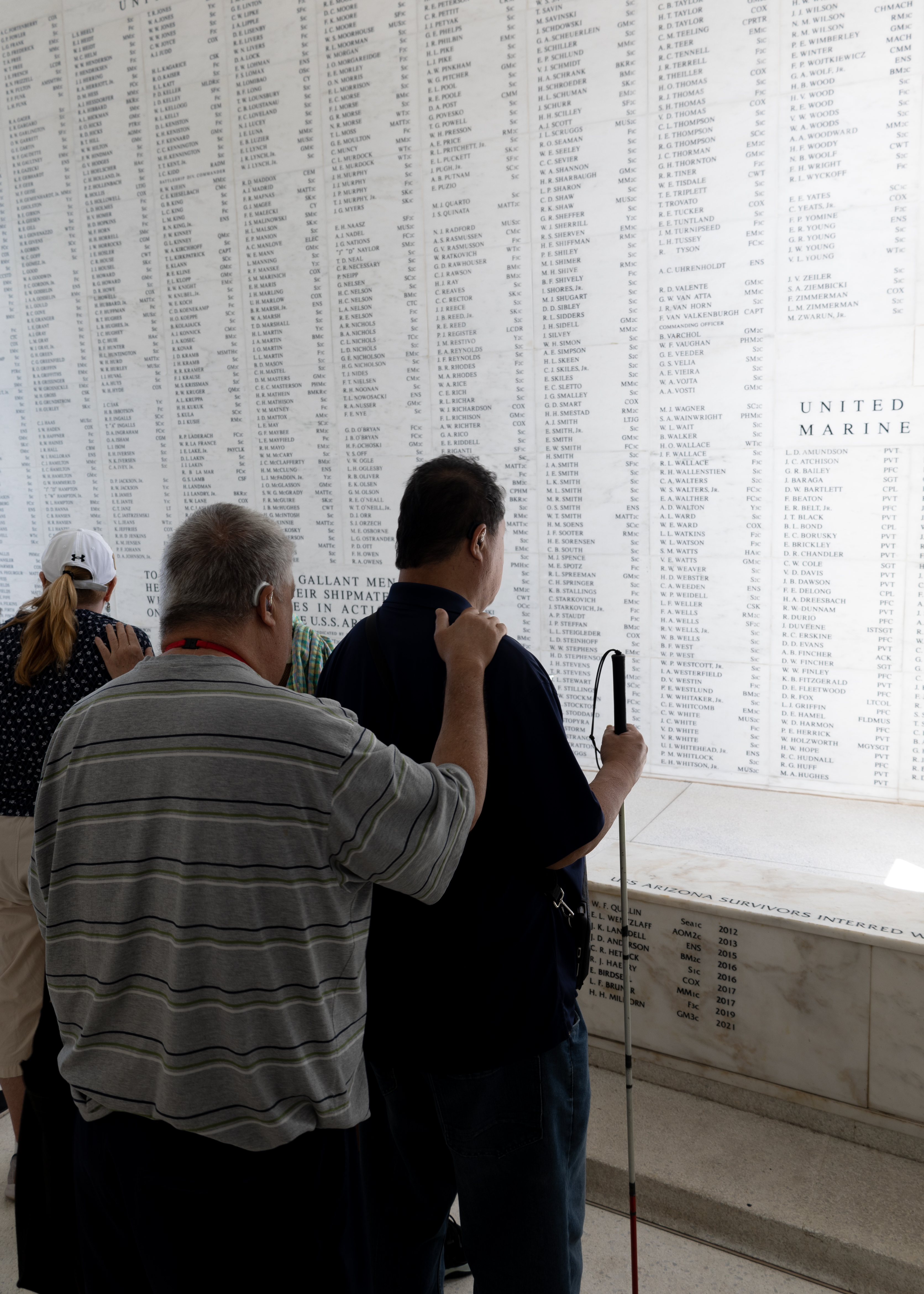 DESCRIBING: A vertical color photograph. DESCRIPTION: Two men, the Kamaka brothers, face a massive marble wall in the U.S.S. Arizona Memorial, with their heads bowed slightly, as in deep thought and contemplation. They are standing in a line, one in front of the other, with the second man putting his right hand on the first man's right shoulder. The wall lists rows and rows of names and military ranks. Those are referring to people who died in the Dec. 7, 1941 attack on Pearl Harbor. Since the men are facing the wall, their faces cannot be seen or described. The man closest to the wall, Lance, is taller than the other, Hilarion, and wearing a dark blue shirt and black pants. The man second in line, Hilarion, is wearing a gray shirt that has horizontal black and white stripes and dark pants. Both have short hair, but the first man's hair color is mostly dark, and the second man's is mostly gray. In front of them is another pair of people also communing with the wall at this site, but they are obscured in the image by the two brothers. Because of that, only a part of one of them can be seen and the back of the other. But through that small window of view, the people can be seen touching, with a hand of the mostly obscured person on the shoulder of the other, a woman in a white hat, with a pony tail. Those two people are looking at the wall but otherwise sharing this space with the Kamaka brothers at the same time and in basically the same ways. CAPTION: Hawaii Association of the Blind members Hilarion and Lance Kamaka stand silently and with reverance near the name-filled marble walls inside the U.S.S. Arizona Memorial. This Pearl Harbor National Memorial site commemorates the nearly 1,200 people who died on that ship when it was attacked and sunk on Dec. 7, 1941 in the Oahu harbor. Hilarion is standing behind his older brother and resting his right hand on Lance's right shoulder. Lance is holding a white cane, and the men also have visible hearing aids, indicating they are both hard of hearing and blind. The brothers recalled visiting the National Park Service site decades ago, as children, but had felt that the site at the time was inaccessible to them and not worthy of a return visit. That changed when they were invited back in April 2023 as part of a research project being conducted by The UniDescription Project. On this return visit, Lance said, the site's accessibility had improved greatly, and this improved access allowed him and his brother to properly pay their respects to the people who had perished and also to learn more about the history of the place and its role in World War II. For our part, the UniD research team has collaborated with the Hawaii Association of the Blind and the Pearl Harbor staff, including Chief of Interpretation David Kilton, to audio describe the site's official print brochures for Pearl Harbor National Memorial, the U.S.S. Arizona Memorial, the U.S.S. Oklahoma Memorial, and the U.S.S. Utah Memorial. Descriptions of those national park sites (and more than 150 others) are available now in the free UniD app (for iOS or Android).